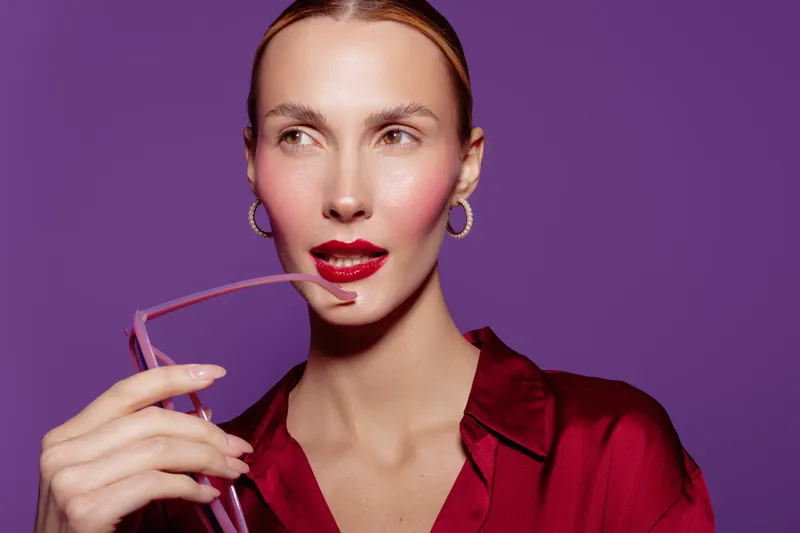 Stylish woman with slicked hair wearing red blouse and hoop earrings holds eyeglasses near lips. Purple background and soft lighting create elegant and modern fashion portrait concept.