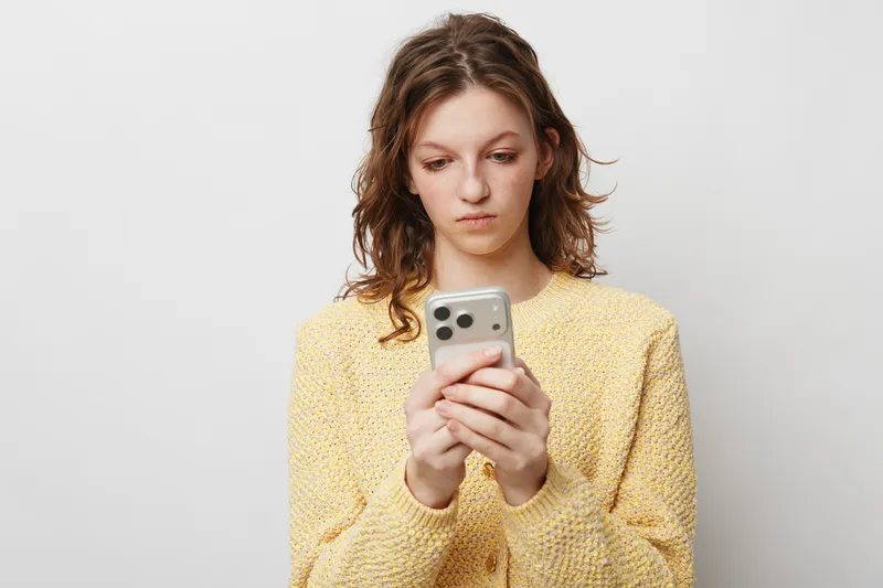 Young woman in yellow sweater looks focused while holding smartphone with both hands on white background. Concept of communication, technology, and social media usage.