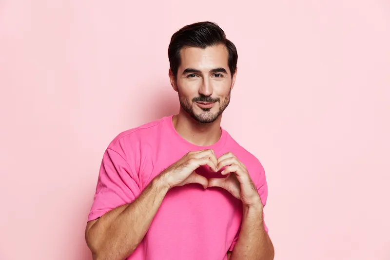 Handsome smiling man in pink t-shirt makes heart shape gesture with hands, looking at the camera against solid pink background. Concept of love and positivity.