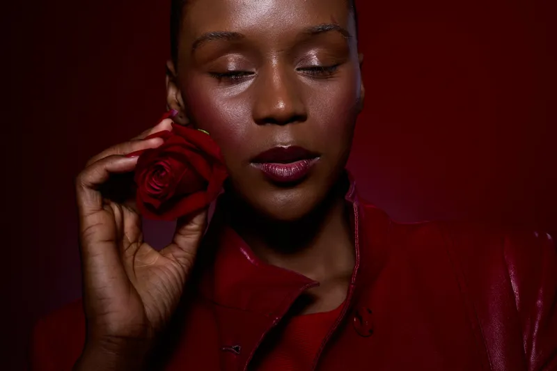Close-up of a dark skinned African woman with closed eyes holding a red rose near her face. She wears red clothing against a dark red background. Concept of beauty and elegance.