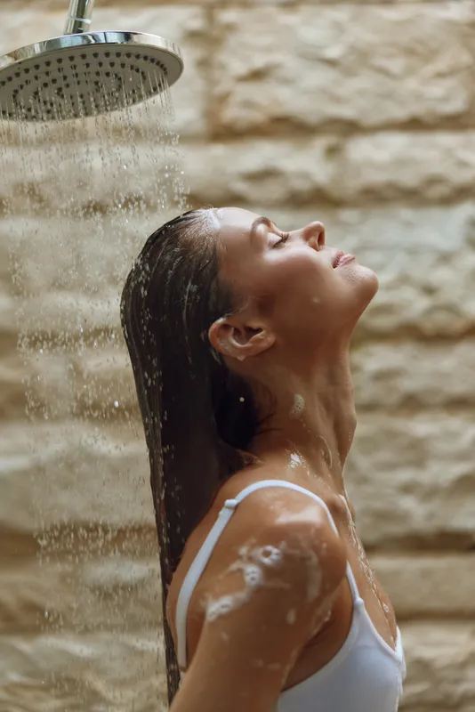 woman showering with closed eyes under warm water spray, relaxing indoors, wet hair, white top, peaceful moment, natural light
