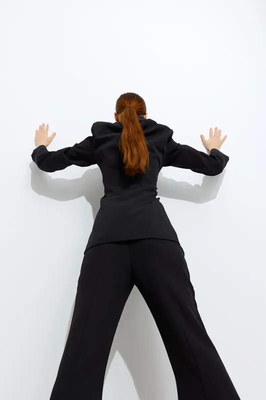 Woman in black business suit with long red hair, standing against white wall with hands pressed, showing professional and determined posture in studio setting.