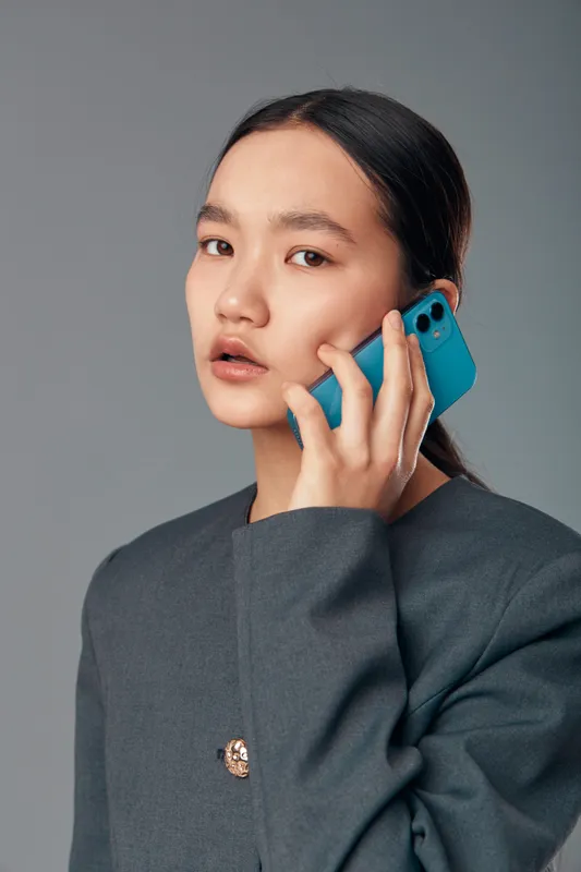 Young woman with serious expression holding smartphone to ear talking on call wearing dark jacket against plain background in studio portrait for communication concept