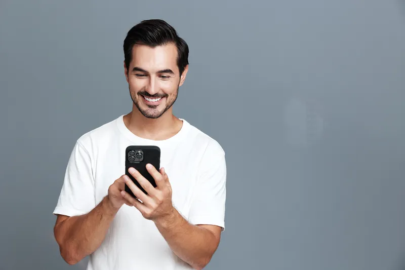 Happy smiling man holding smartphone, dressed in casual white t-shirt, having fun while browsing the web, isolated on solid gray background, studio portrait.