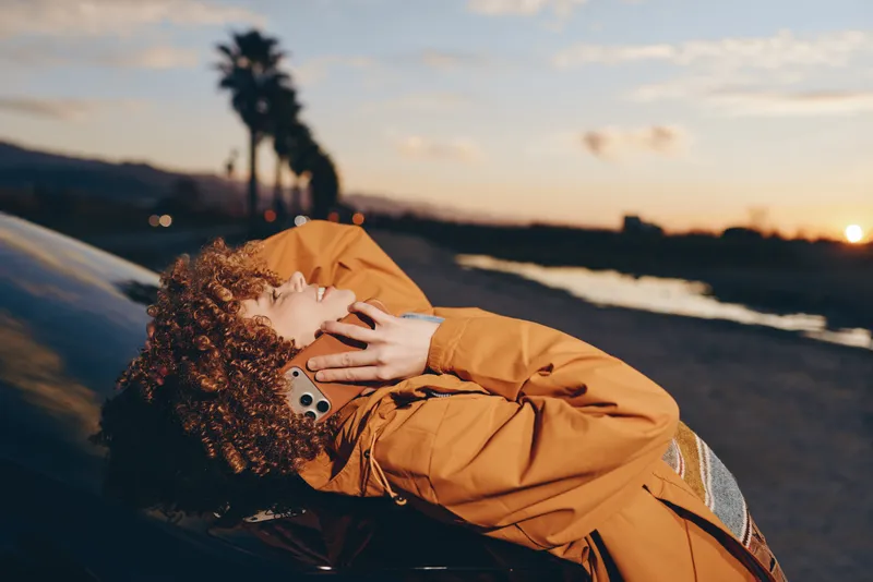 woman lifestyle on beach at sunset, curly hair in orange jacket, holding smartphone near face, relaxed outdoor mood, golden sky glow, travel feeling and personal freedom, scenic coast