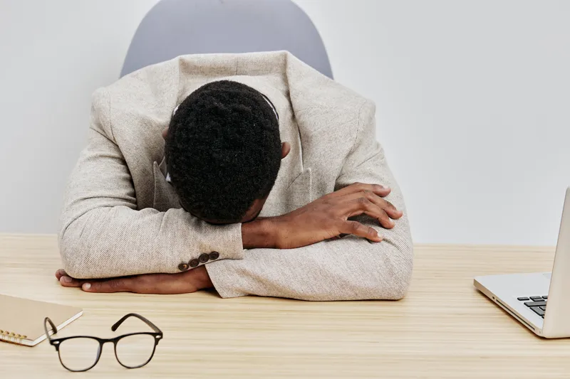 Overwhelmed young African American man wearing beige sweater sitting at desk with head resting on arms, feeling stressed or exhausted in modern office environment, business concept, black and white im