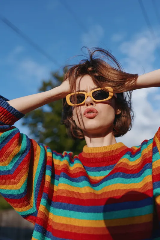 Young woman in rainbow sweater and yellow sunglasses outdoor portrait, short brown hair, hands in hair, playful expression on sunny day, urban lifestyle fashion portrait for social media.