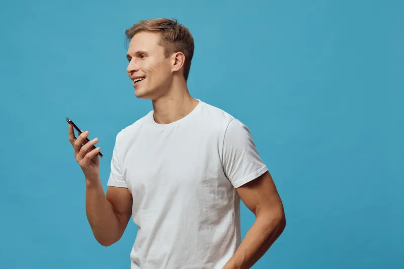 Smiling young man with short blond hair in casual white t-shirt holding smartphone in hand against bright blue background, looking away and enjoying technology use, portrait shot, lifestyle concept, v