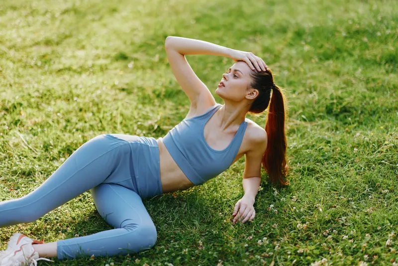 Active young woman in sportswear lounging on grass, showcasing a healthy lifestyle, fitness, and mindfulness in a vibrant green outdoor setting