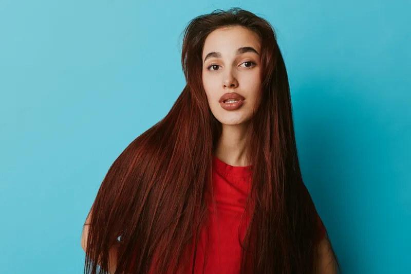 Young woman with long straight hair and natural makeup wearing red shirt posing against blue background. Beauty, fashion, and casual style with confident expression and healthy hair.