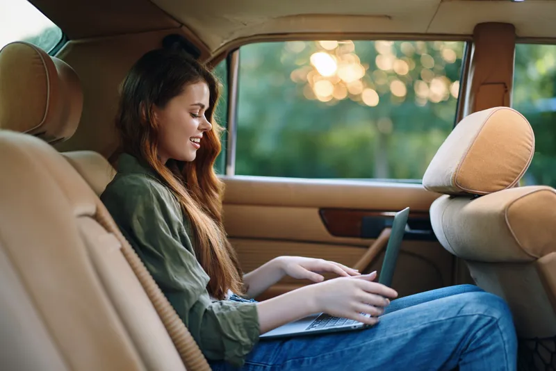 Young woman working on a laptop in a car during sunset, wearing casual clothes and smiling, highlighting a sense of productivity and relaxation.