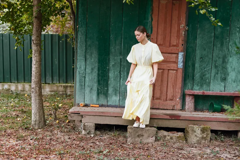 Woman wearing a pale yellow dress stands on a rustic porch outdoors, surrounded by greenery and fallen leaves, creating a calm fashion moment in natural light and peaceful scenery.