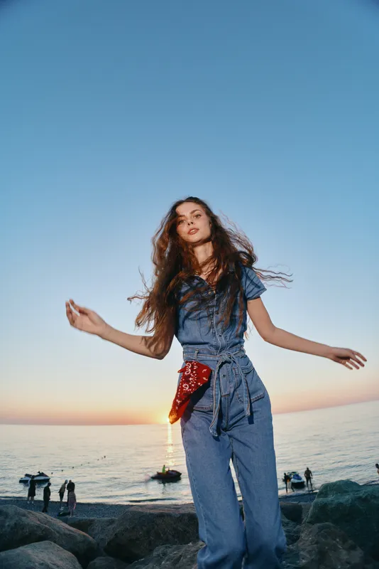 young woman dancing at sunset on the beach wearing denim outfit with flowing hair and ocean background in warm natural light