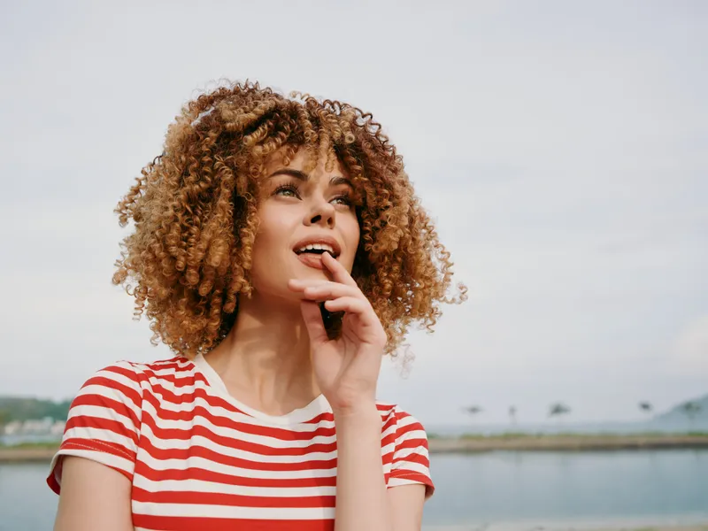 Close up portrait of a young woman with curly hair wearing a red striped shirt, standing outdoors near water with a calm sky, thoughtful expression, and hand near her lips.