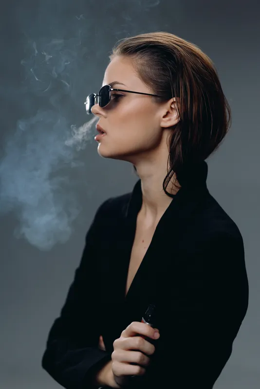Stylish woman wearing sunglasses smoking vape with smoke against dark background