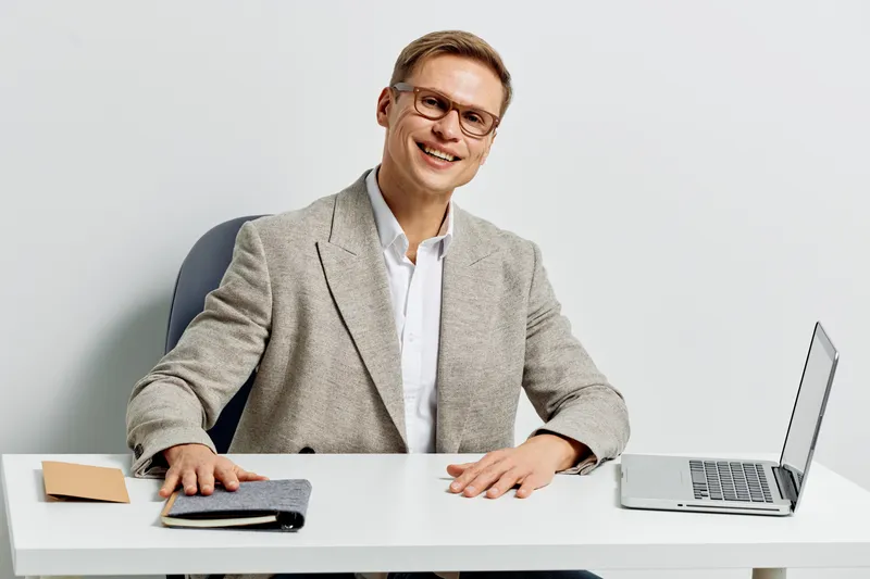 Smiling man in beige blazer sitting at desk with open laptop, notebook, and pen in modern office. An approachable professional looking at camera, cheerful confident expression. People lifestyle concep