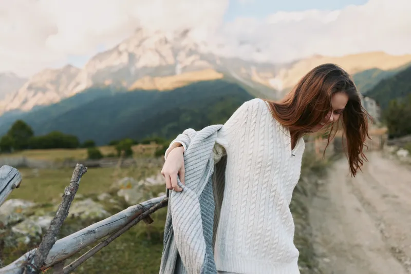 Young woman with long brown hair walks along a rural dirt road wearing a white knitted sweater while holding a gray scarf in a mountainous landscape during daylight.
