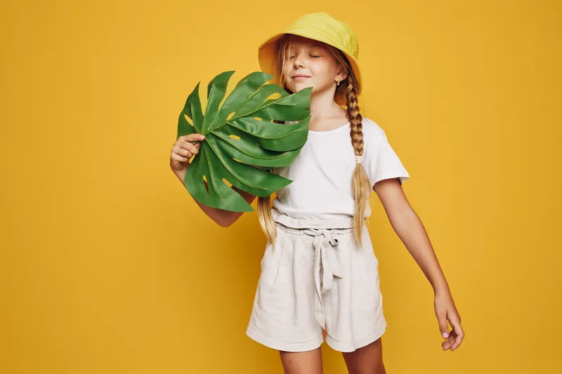 Young girl with braided hair wears a yellow hat and holds a large tropical leaf on a bright yellow background.
