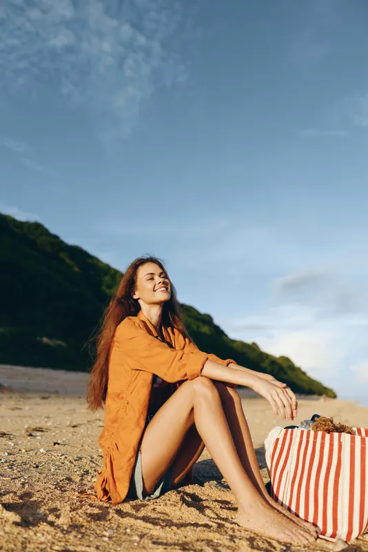 Young woman with long brown hair sits on the sandy beach in an orange dress, smiling as a striped bag rests beside her, with legs bent and a warm sunlit glow on the hillside.