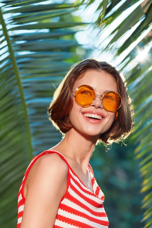 A cheerful woman wearing a red and white striped top and orange sunglasses smiles in bright sunlight, framed by tropical leaves, creating a lively, sunny summer vibe.