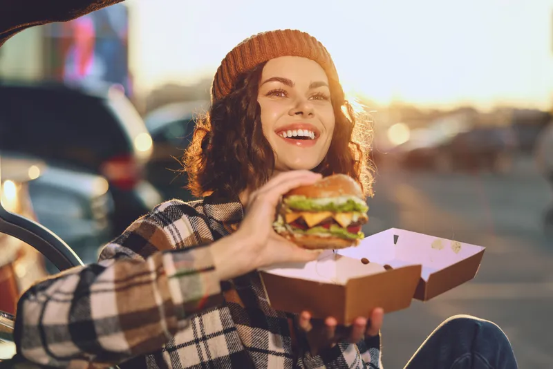 Woman holding burger and takeaway box, smile radiant in golden hour glow of an urban street, candid lifestyle portrait with beanie and plaid coat, authenticity, emotional storytelling, mindful living.