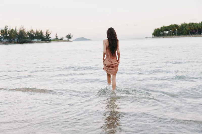 A tranquil seaside scene features a lone figure wading into shallow water, soft light, calm waves, and a distant tree line along a quiet horizon, in distance.