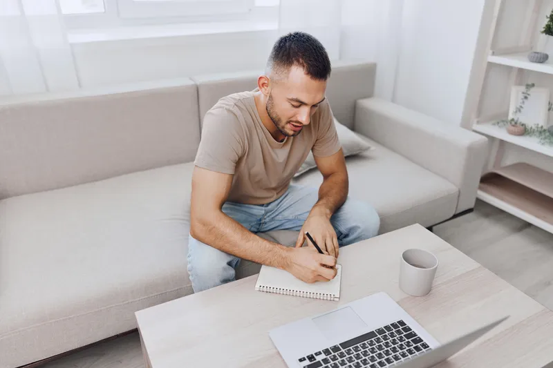 Focused man writing in a notebook at home, reflecting on work-life balance and mental health, while surrounded by a calming environment for personal growth.