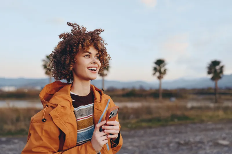 Woman lifestyle outdoors, smiling with curly hair, orange jacket, holding coffee cup, soft daylight, blurred palms and road in background, casual travel mood, warm sunny morning