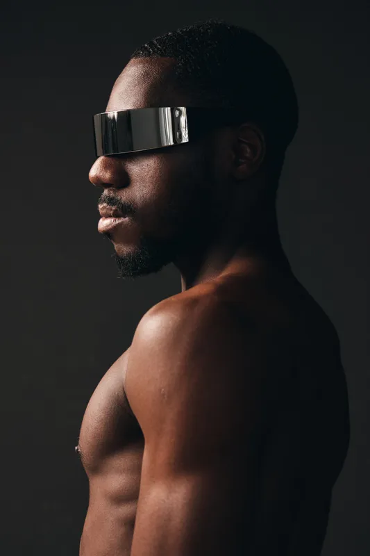 Creative visual designer style portrait of black man with dark skin wearing futuristic reflective visor glasses in studio. Side view, bare torso and modern fashion concept with minimal lighting.