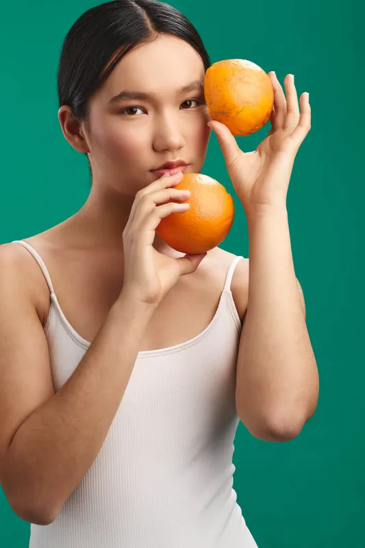 asian woman holding oranges in a studio setting with a green backdrop. She gazes at the camera with a soft expression, presenting a fresh and vibrant fruit portrait.