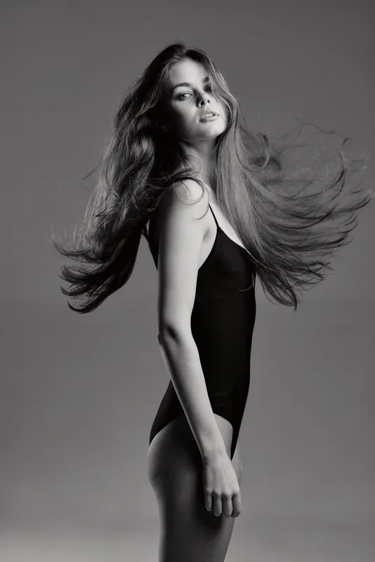 Black and white portrait of a young woman with flowing hair in a black bodysuit, showing elegance and motion on a plain background.