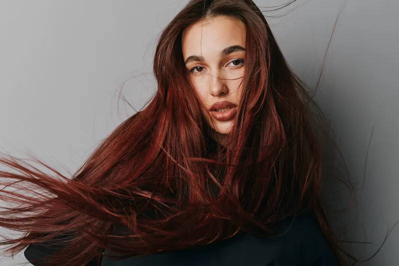 Woman with long red hair and natural makeup posing in studio with gray background. Closeup portrait of young female with flowing hair and serious expression wearing dark top.