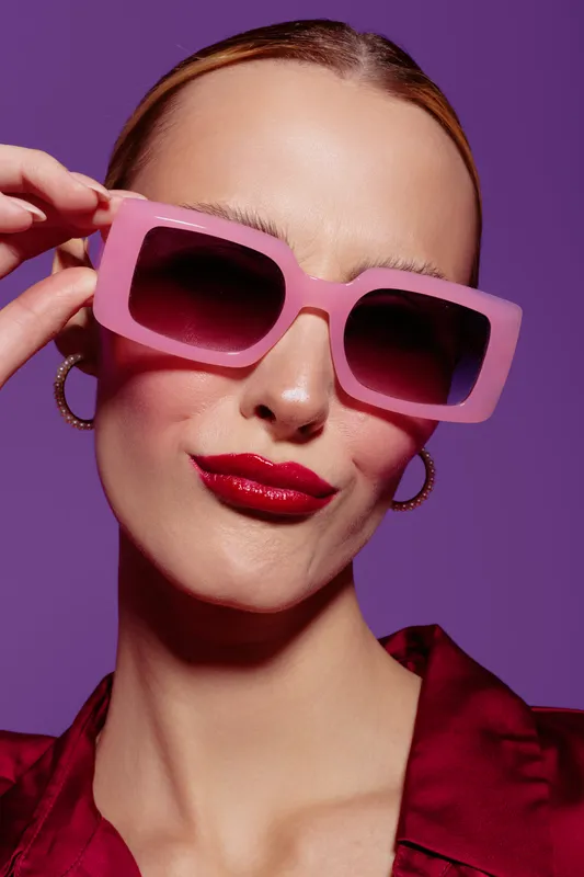 Stylish woman with pink sunglasses and red lipstick poses confidently against purple background. Fashion concept with bold accessories and vibrant colors.