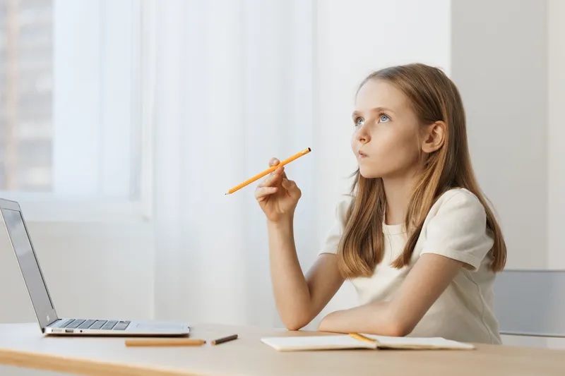 Thoughtful young girl pondering ideas while studying at a desk with a laptop, pencil in hand, in a bright and minimalistic room