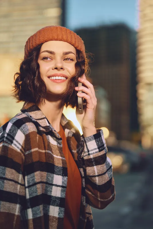 Woman on phone smiling in city street at golden hour with beanie and plaid jacket, outdoor portrait in warm sunlight capturing authenticity, candid lifestyle, mindful living and emotional