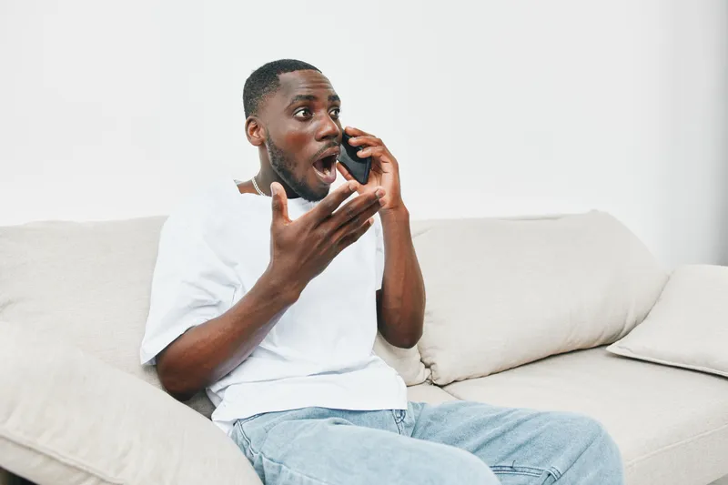Shocked African man talking on the phone while sitting on the couch, expressing surprise and excitement in casual home setting, light color palette