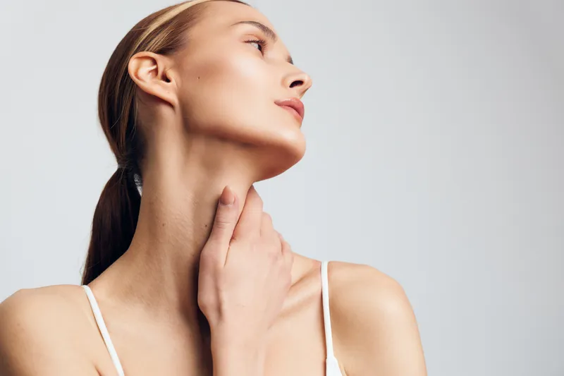 Young woman with clear skin and blonde hair touches her neck and looks sideways with calm expression, isolated on white background in minimalistic style portrait.