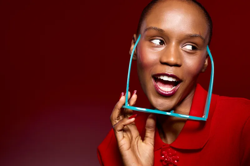 Happy African American woman with dark skin wearing red coat holds blue sunglasses near face smiling on red background. Concept of fashion, beauty, and joyful lifestyle portrait.
