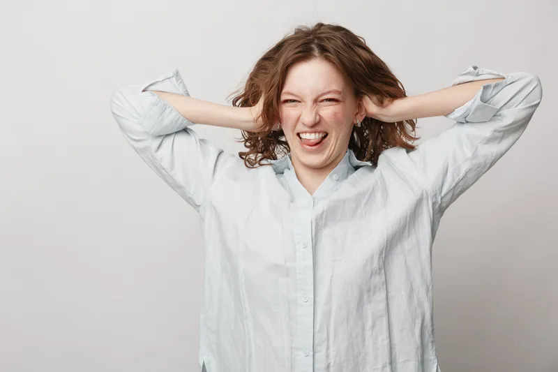 Young woman in white shirt makes funny face with tongue out and hands behind head. Studio portrait on light background. Concept of humor, playfulness, and carefree attitude.