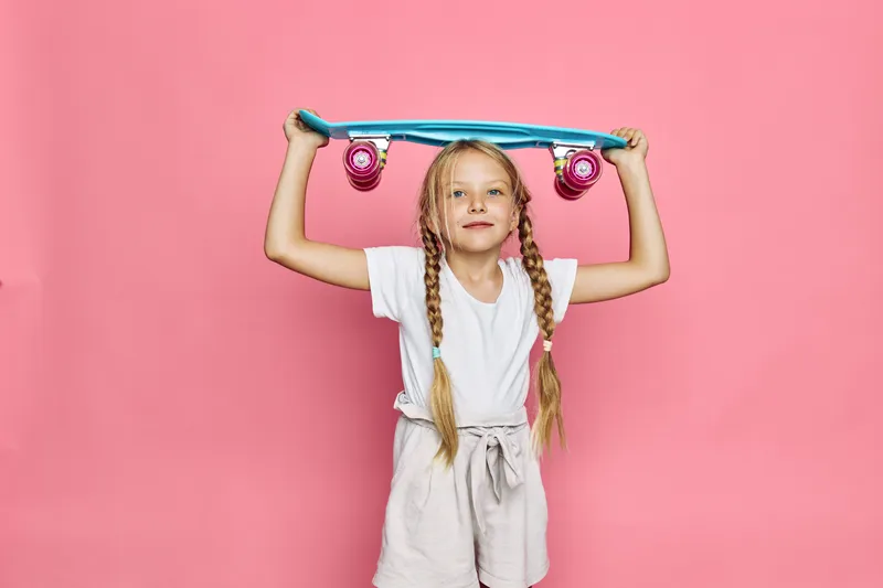 Young girl with braids holding a blue skateboard above her head smiling. Studio shot on a pink background. Concept of childhood, leisure, and fun.