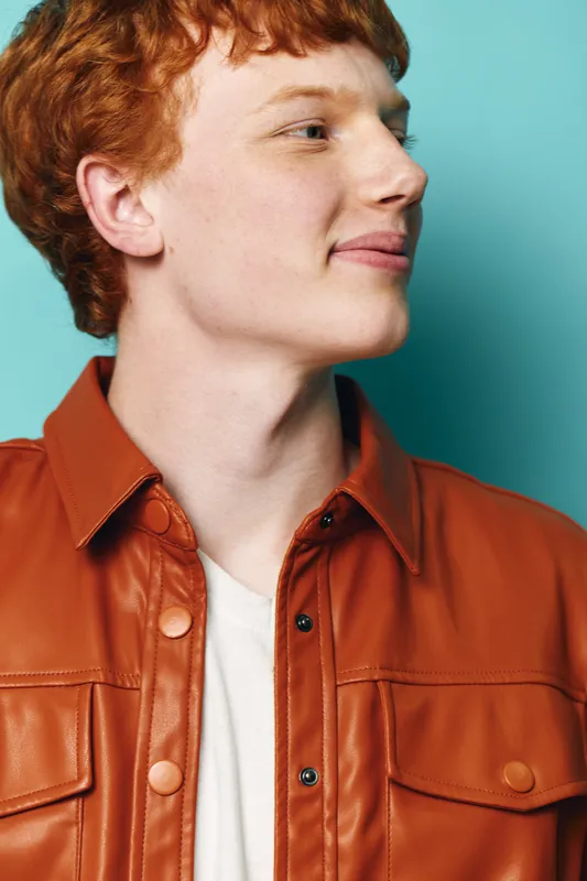 Young man with red hair wearing brown leather jacket and white shirt, looking to side with slight smile on blue background. Casual style and confident pose portrait.