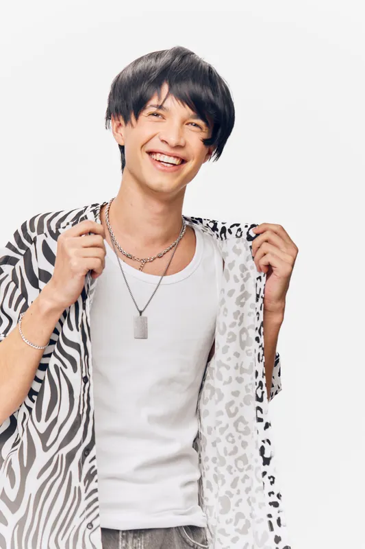 smiling man wearing patterned jacket and white tank top with layered necklaces, standing against plain white background, youthful and casual style, happy expression