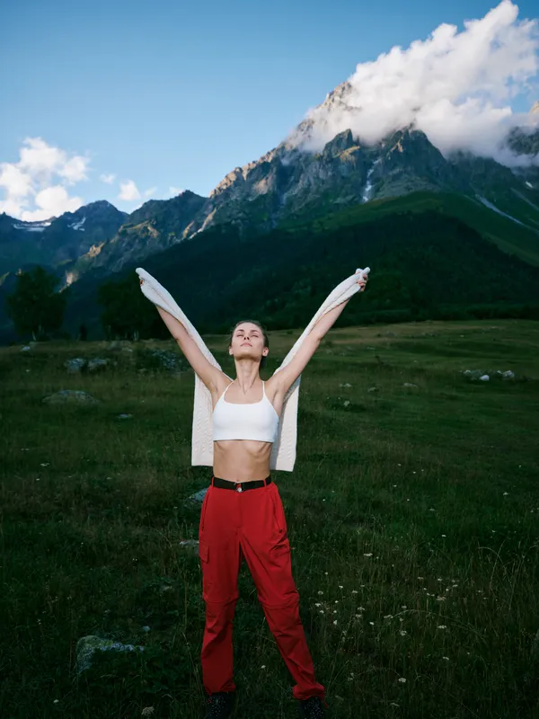 Outdoor scene with a athletic woman in a white crop top and red pants, arms raised, standing in a meadow under rugged mountains and blue sky, conveying energy, freedom, and outdoor fitness