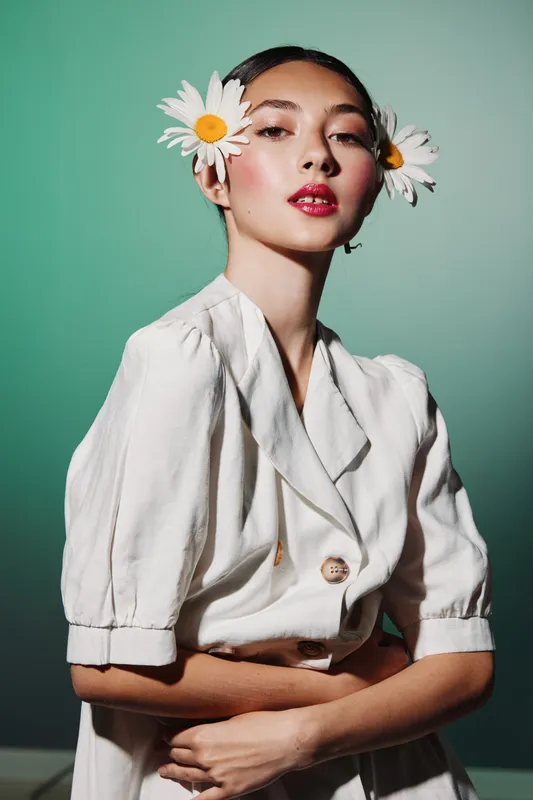 Fashionable woman with two daisies in her hair posing in a stylish white dress on green background. Her clean makeup and confident expression highlight creative beauty concept.