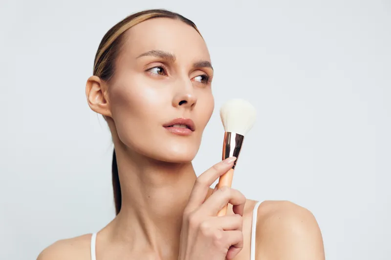 Young woman holds makeup brush near face looks away with clean skin and natural makeup isolated on white background beauty concept skincare routine.