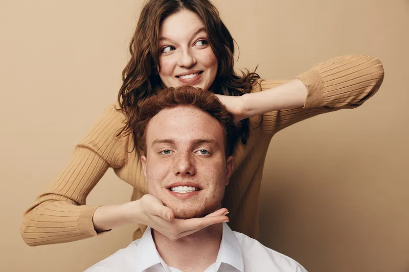 Smiling young woman with brown hair playfully framing the face of a happy redhead man in white shirt. Warm beige background creates cozy and friendly atmosphere. Concept of friendship and joy.