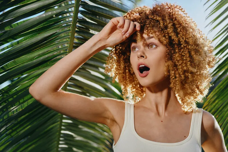 Young woman with curly hair wearing a white tank top looks surprised while standing near tropical palm leaves under bright sunlight.
