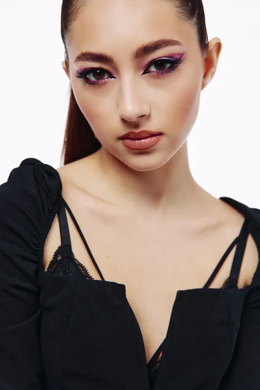 Close-up portrait of a young woman with edgy purple eye makeup and nude lips wearing a black top with visible straps. Studio shot on a white background. Beauty fashion creative concept.