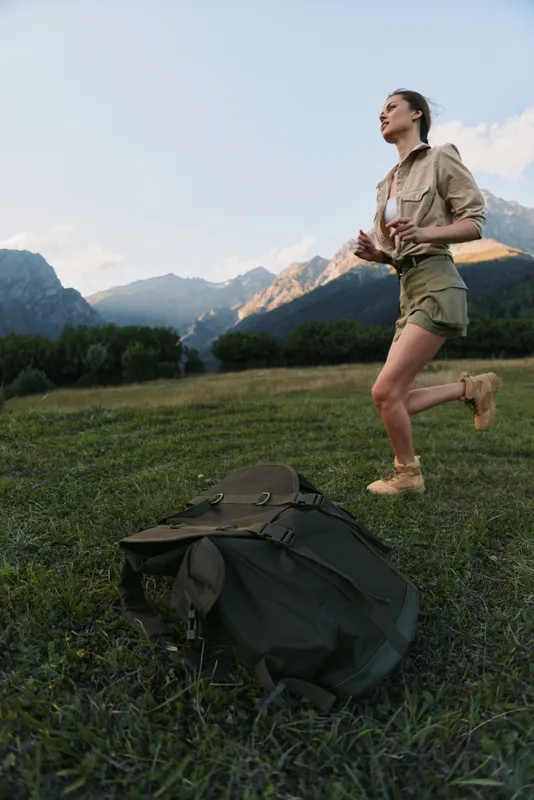 A young woman runs across a grassy field with a backpack near rugged mountains, conveying an adventurous hiking moment, energy and freedom