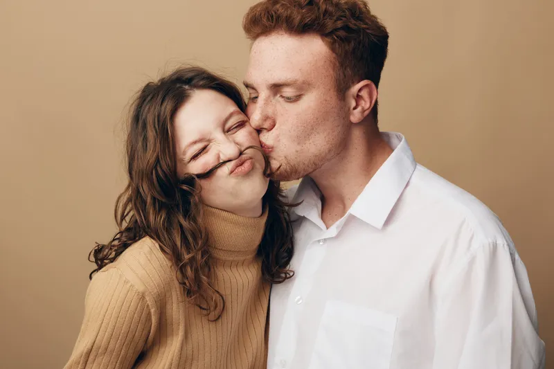 Young couple in casual clothes share a tender moment as the man kisses the woman on the cheek. Warm beige background highlights their affectionate and playful relationship.